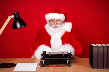 Santa at the desk typing on a vintage typewriter in a festive red studio with lamp and books...