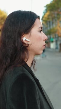 Self-assured young professional with flowing dark hair walking a busy city street, enjoying music or a podcast on wireless earbuds during her urban commute to work, stylish and confident