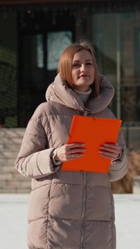 Seasoned real estate agent presents a modern home on a sunny winter day, holding an orange folder and smiling confidently at the camera while inviting buyers to tour the property