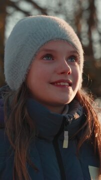 Happy little girl with blue eyes looking up with a hopeful and dreamy expression, illuminated by the warm light of the setting sun in a winter park. Her face shows pure joy and wonder