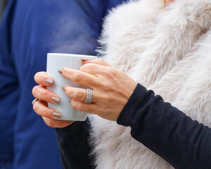 A visitor at the autumn Naplavka market holds a warm drink in a white mug, hands adorned with glittery nails and rings, capturing the cozy atmosphere and seasonal charm of this popular riverside event