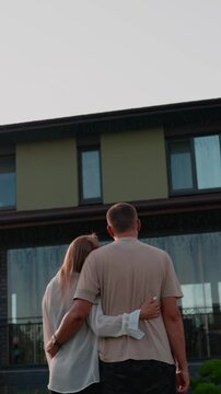 Young couple embraces in the rain, gazing at their contemporary house, representing their joint aspirations, investment in real estate, and promising future in their new home