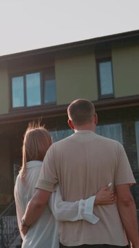 Young couple embracing in their backyard, gazing at their modern suburban home. The new homeowners are seen from behind, looking ahead to their shared future with excitement and love
