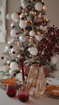 Elegant holiday table with red berry accents, candles and dried orange slices on plates, while a softly blurred, glowing christmas tree creates a cozy festive atmosphere