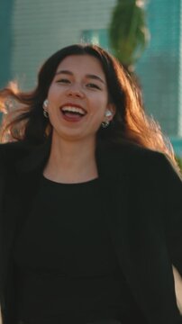 Joyful young businesswoman with wireless earbuds dancing happily in a modern city at sunset, her long hair blowing in the wind as she celebrates success with a radiant smile