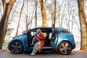 Side view, with cup of drink. Beautiful blonde woman is with her blue electric car on the road of forest
