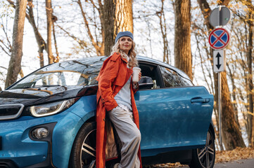 Leaning on the automobile, holding cup of drink. Beautiful blonde woman is with her blue electric car on the road of forest