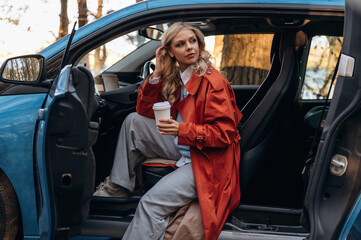 Side view, with cup of drink. Beautiful blonde woman is with her blue electric car on the road of forest