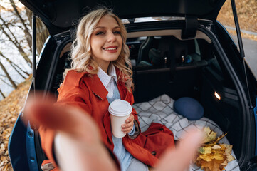 Selfie time. Positive facial expression. Beautiful blonde woman is with her blue electric car on the road of forest