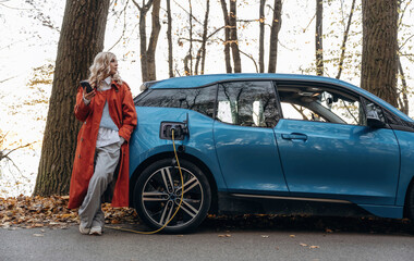 Automobile is charging. Beautiful blonde woman is with her blue electric car on the road of forest