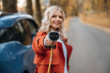 Holding the charging cable. Beautiful blonde woman is with her blue electric car on the road of forest