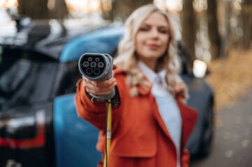 Holding the charging cable. Beautiful blonde woman is with her blue electric car on the road of forest