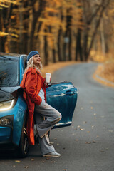 Smiling, having fun. Beautiful blonde woman is with her blue electric car on the road of forest
