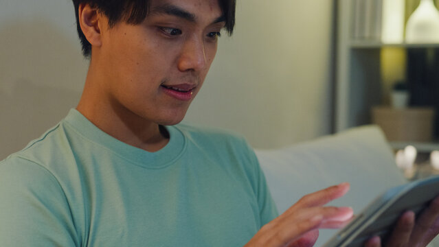 Close up young Asia male with casual cloth sit on sofa finger scroll tablet watch online movie in living room cozy house at night. E-book reading, Shopping online, Lifestyle leisure at home concept.