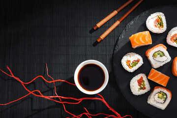 A salmon maki rolls with vegetable and cheese fillings on black slate plate, chopsticks, soy sauce bowl and red wavy branches on black bamboo mat background.