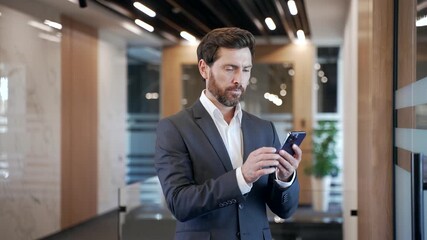 Businessman in formal suit is using browsing mobile phone while standing in hallway of a modern business center. Confident handsome manager is chatting online or reading writing message on smartphone - Powered by Adobe