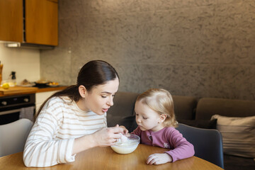 Naklejka premium Happy single mother doing her daughter's hair while having breakfast. Woman combs the hair of a little girl. The concept of morning routine, single parenthood and preparation for school