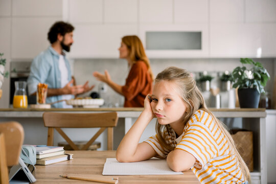 Bored child sitting at table with parents arguing in background