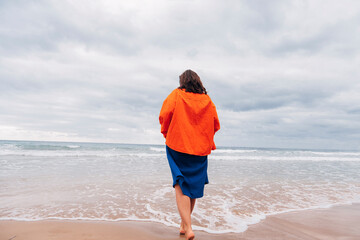 Woman in orange jacket walking barefoot on sandy beach by the sea