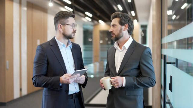 Two businessmen in formal suits engaging in casual discussion standing in the corridor of modern business center. Colleagues have friendly conversation during break, talking, sharing ideas with coffee
