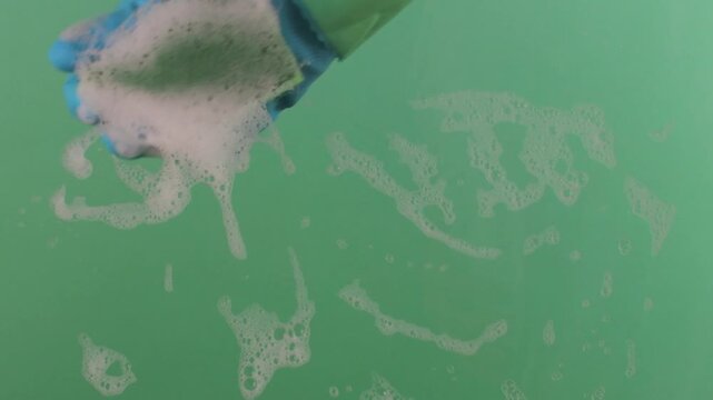 A hand in a rubber glove with a sponge washes the surface. Hands in rubber gloves hold the sponge and scrub the surface. The sponge is covered in sudsy cleanser on a chroma key.
