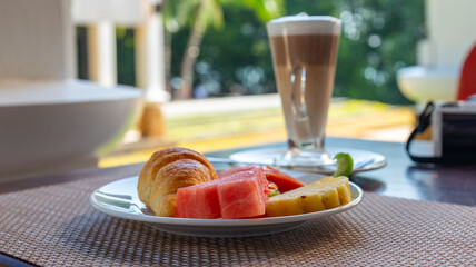 Tropical resort breakfast: Croissant, fresh watermelon, and pineapple served on a white plate with a tall glass of layered latte on an outdoor patio with a green, tropical background.