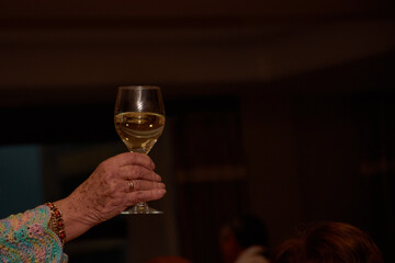 An elderly person's hand raises a glass of white wine in a toast during an intimate celebration against an elegant black background.