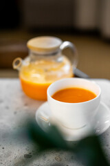A close-up shot of vibrant orange carrot or pumpkin soup served in a white cup and saucer, with a matching glass teapot in the blurred background on a modern concrete table.