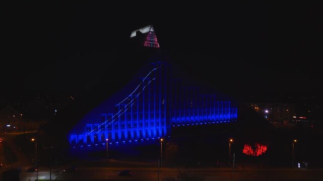 Aerial night view shows Latvian National Library in Riga during Riga Staro 2025. Blue LED wash, light mapping, pulsing crown, red heart lights, cars and street lamps.