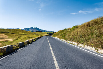 Empty asphalt road winding through the rolling grassy hills with a distant mountain range on a sunny day.