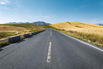 Empty asphalt road leading towards the rolling golden hills and magnificent mountain range under a clear blue sky.
