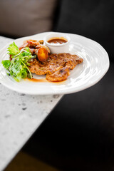 A delicious, cooked meat cutlet, likely a pork chop, served on a white plate with roasted potatoes, a green salad garnish, and a side of brown sauce in a restaurant setting