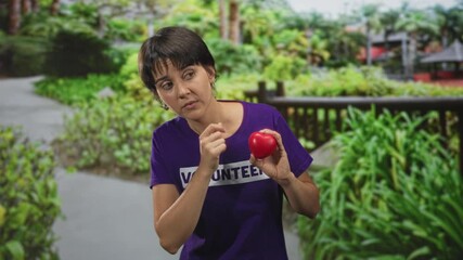 Woman volunteer in purple shirt holding red heart, hand to chin pensive pose on park path in forest; compassion reflection.