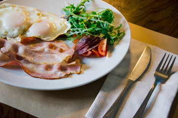Classic breakfast plate featuring fried eggs, crispy bacon, fresh greens, and sliced tomatoes served on a white dish.