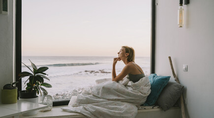 Thoughtful woman relaxes by the window. Woman sitting by panoramic window with ocean view, wrapped in blanket enjoying morning. Serene female in blanket sits on the windowsill with sea view.