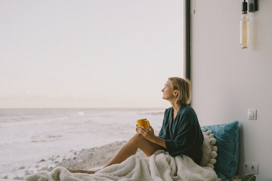 Thoughtful woman with a cup relaxes by the window enjoying morning. Beautiful woman in blanket sits on the windowsill by panoramic window with ocean view enjoying coffee or tea.