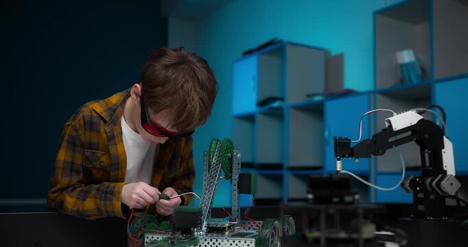 A smart Caucasian boy with glasses is focused on building a robot car. he connects the wires at work. the concept of stem education