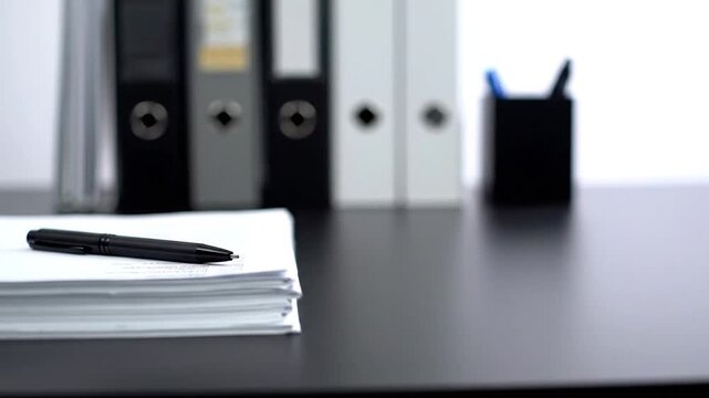 Desk with a pen and a stack of papers in focus; blurred binders and pen holder in background