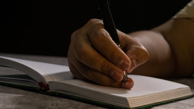 Hand holding a black pen over an open notebook, captured with dramatic shadows. Sharp focus on fingers and pen tip conveys concentration and detailed writing work.