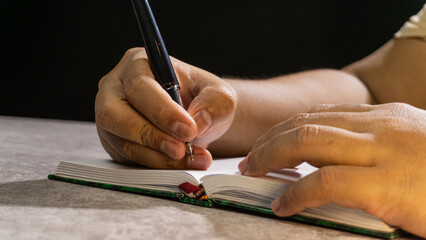 Hands writing in a small notebook under warm lighting. The pen tip and lightly curved pages create...