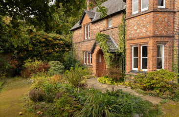 Obraz premium A quaint brick cottage surrounded by lush greenery, ivy-covered walls, and a wooden picket fence. Warm, inviting garden with mature shrubs creating a cozy, rural home atmosphere.