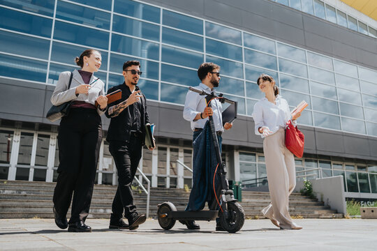 Four business colleagues discuss work while walking near a contemporary office complex. One colleague holds an electric scooter, suggesting active and eco-friendly transportation. - Powered by Adobe