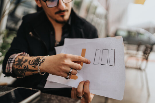Close-up of a man holding papers with sketches, presenting ideas in an outdoor setting. Details include hand tattoos, stylish accessories, and a focus on collaboration and creativity.