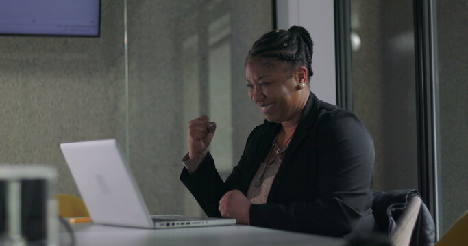 African American businesswoman celebrating success with joyful fist pump while seated at laptop in office meeting room - Powered by Adobe
