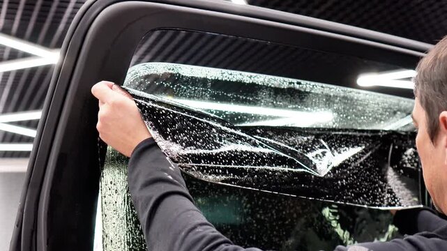 Applying tinting foil on a car window in the tuning studio. Male car service worker glue window tinting in luxury car detailing car repair the concept of love for cars and protection. Wrapping auto. 