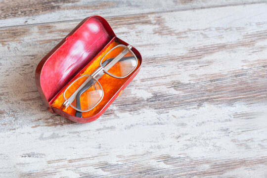 Old glasses with metal frame and black temples lying in a vintage bright burgundy plastic case in daylight on a wooden background in a minimalist vintage context from the 60s and 70s, close-up.