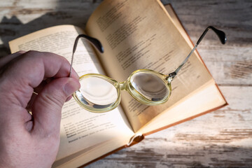 Hand holding reading glasses with high optical power lenses for nearsightedness above a book in bright sunlight, close-up.
