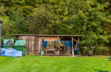 A rustic wooden shed houses hanging nets, tools, and various outdoor equipment. Surrounded by bright green grass and forest trees, the scene conveys utility, craftsmanship, and rural work life.