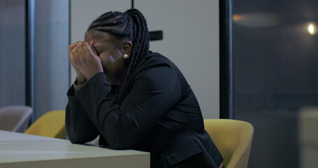 African American businesswoman sits with face buried in hands at office desk, struggling with...
