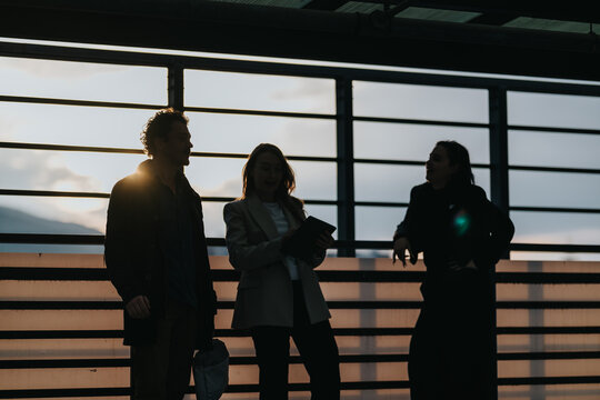 Three people stand on a balcony at sunset, their silhouettes framed by railings. One holds a tablet while the others chat, creating a relaxed, urban moment of connection and camaraderie.